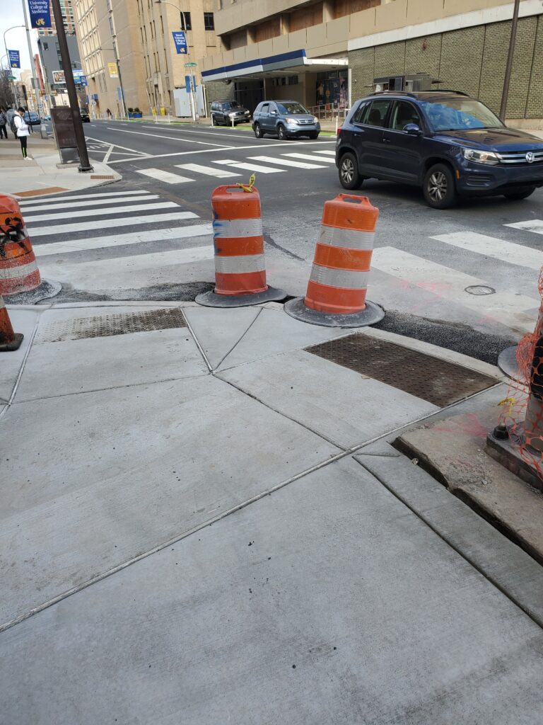 Image of a street corner with ramps in both directions. Large caution barrels are still in place between the ramps. Cars drive by on the street.