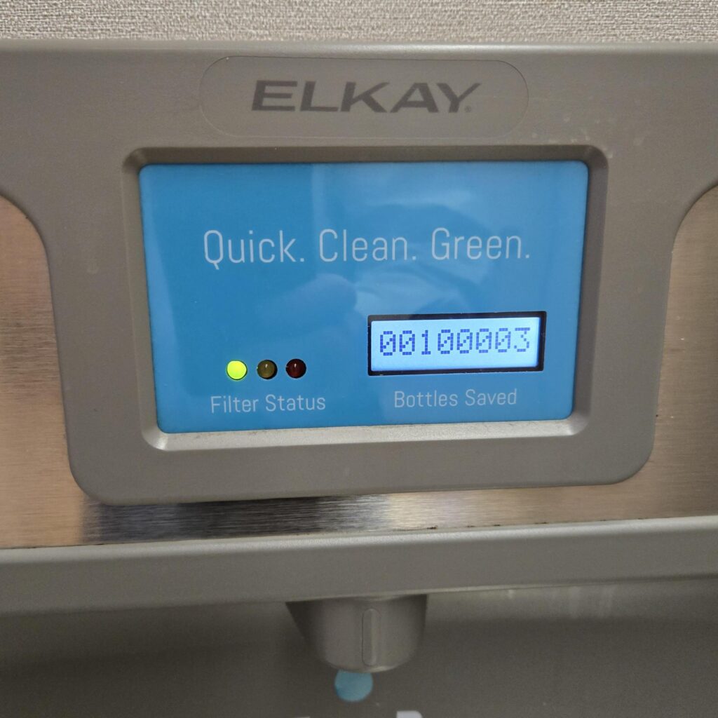 Close-up view of the top of a water fountain with a bottle filler. Text says Elkay. Quick. Clean. Green. Filter status shows a green light. The digital meter reads 100,003.