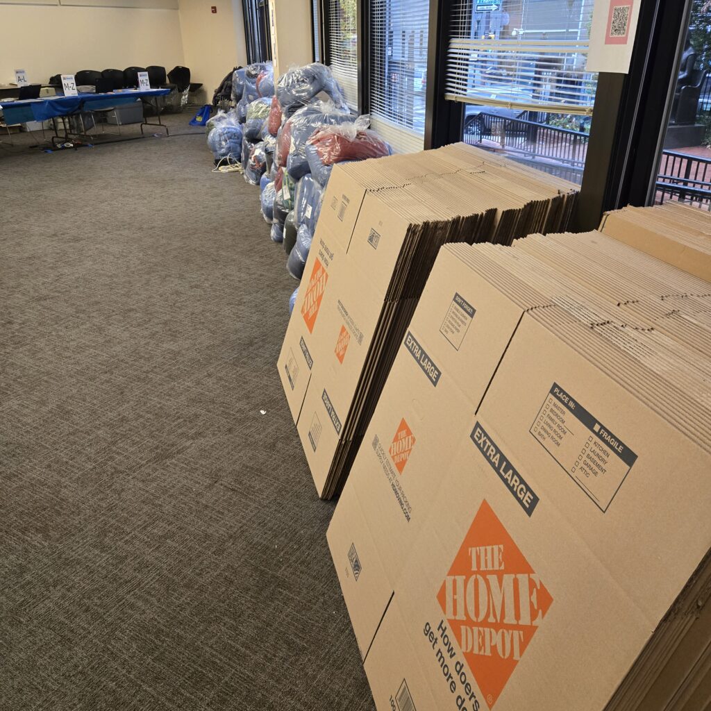 Two stacks of extra large moving boxes are in the foreground. They say Home Depot on them. Behind them is a stack of sleeping bags up against a low wall with tall windows. In the background on the left are a table and some chairs.