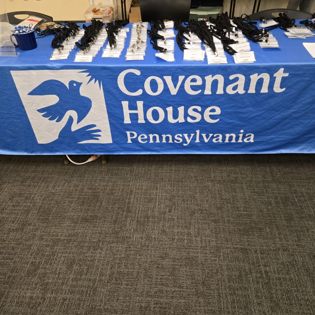 A blue tablecloth says Covenant House Pennsylvania in white letters. A logo on the left shows a dove flying up from a hand toward the sun. On top of the table are columns of neatly arranged name tags. They are for the participants of the Covenant House Sleep Out at Friends Center.