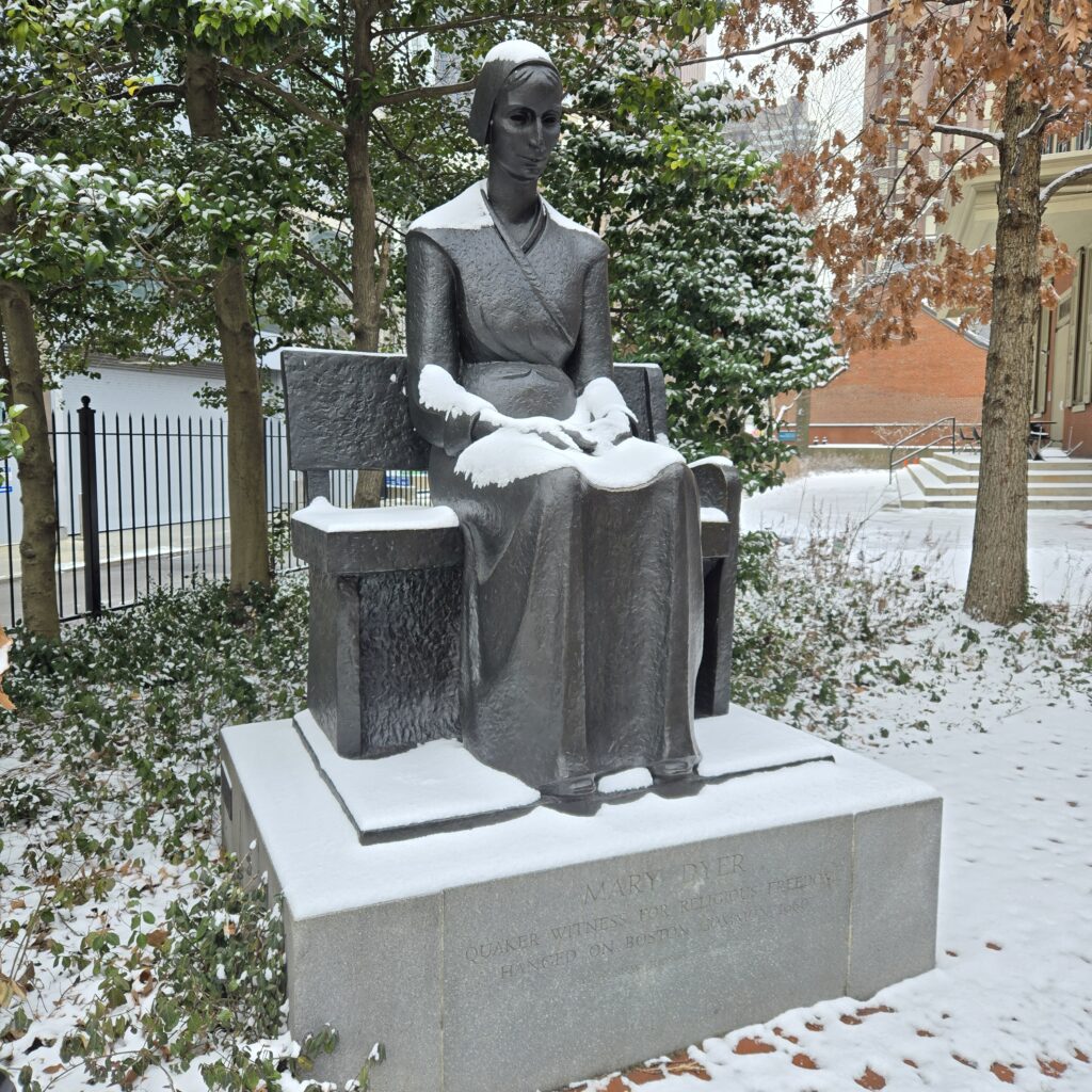 A statue of Mary Dyer. She is sitting on a bench with her hands on her lap. She has snow on her lap, forearms, shoulders, and head. The statue is set on a raised stone plinth. On the left and behind it are holly trees. On the right is an oak. Behind that tree are brick buildings, including the porch of the Race Street Quaker Meetinghouse.