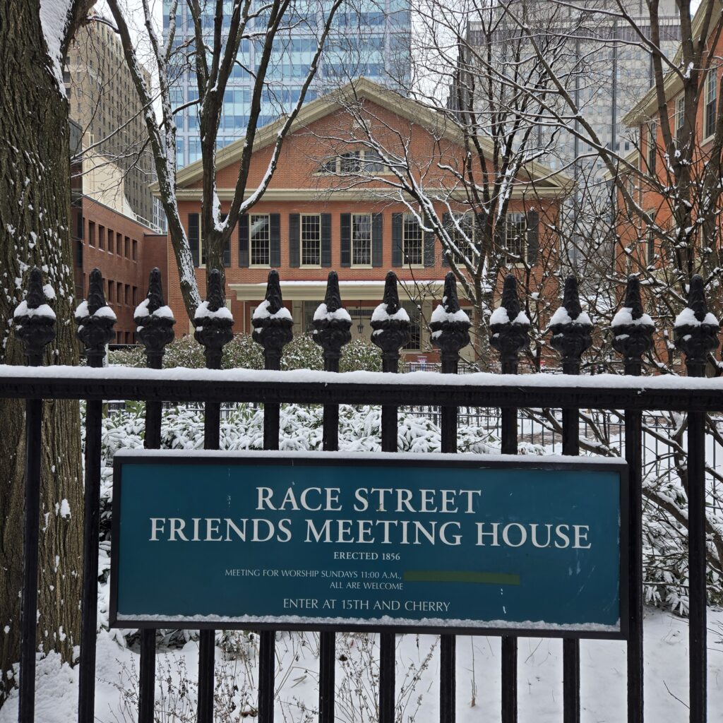 A sign on a black, old-fashioned fence says Race Street Friends Meeting House. Erected 1856. Enter at 15th and Cherry. Behind the fence are some trees, and beyond them is the Race Street Friends Meetinghouse. We usually close it up and make it one word. Other buildings are in the background. Here, Friends refers to the Religious Society of Friends, also known as the Quakers.