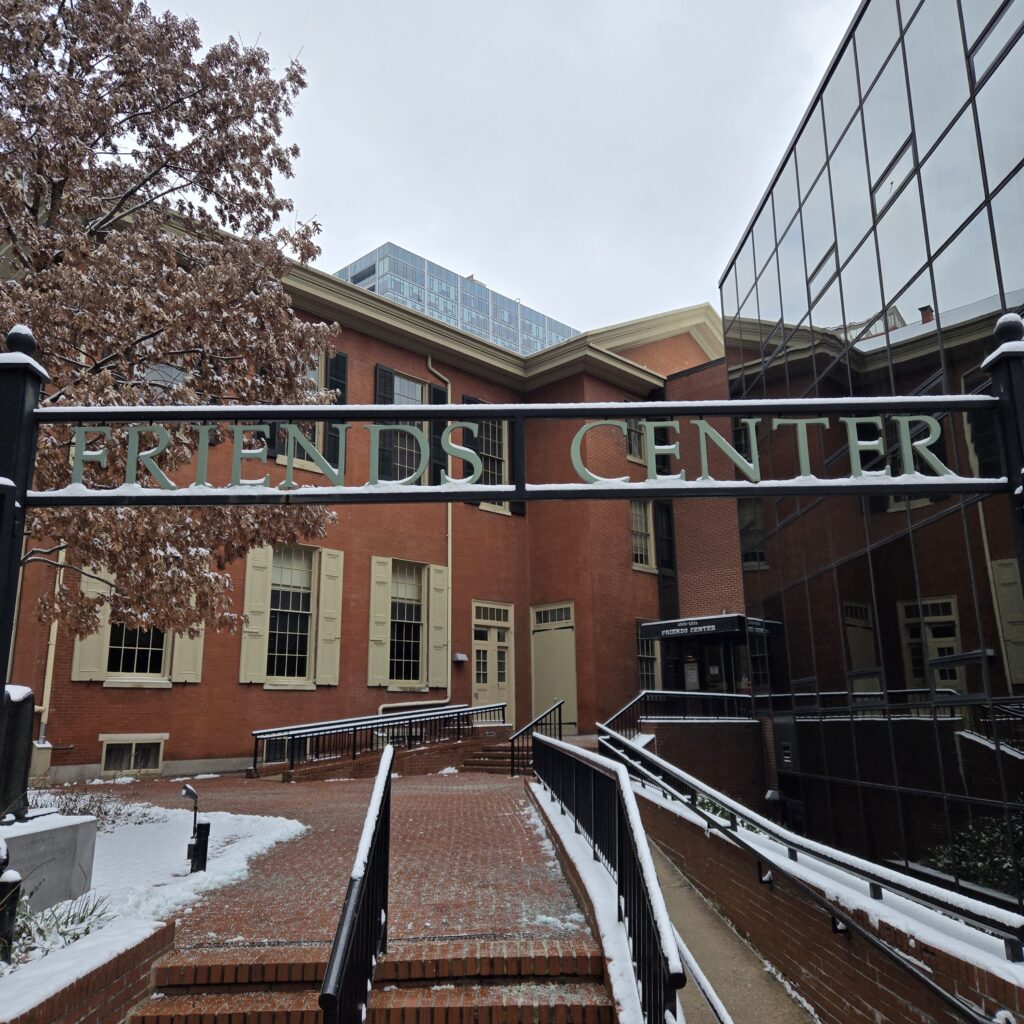 A sign across the top of an open gate says Friends Center. On the right side is a glass curtain wall of a nonprofit office building. Ahead is a brick two story building with beige shutters and trim on the first floor and black shutters on the second floor. It is the Race Street Quaker Meetinghouse. A tall modern building is behind it in the distance. A white oak tree that still has brown leaves on it is behind the gate to the left. Bits of snow are clinging to the leaves. In the top center the sky is gray and overcast.