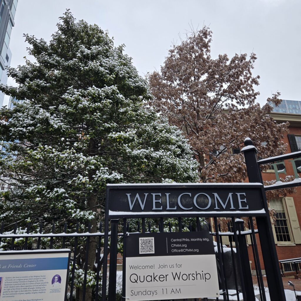 Snow-covered holly and oak trees are in front of a brick building. A sign on top of the fence says Welcome. A sign attached below it says Welcome! Join us for Quaker worship Sundays at 11 am. Yes, this welcome includes you.