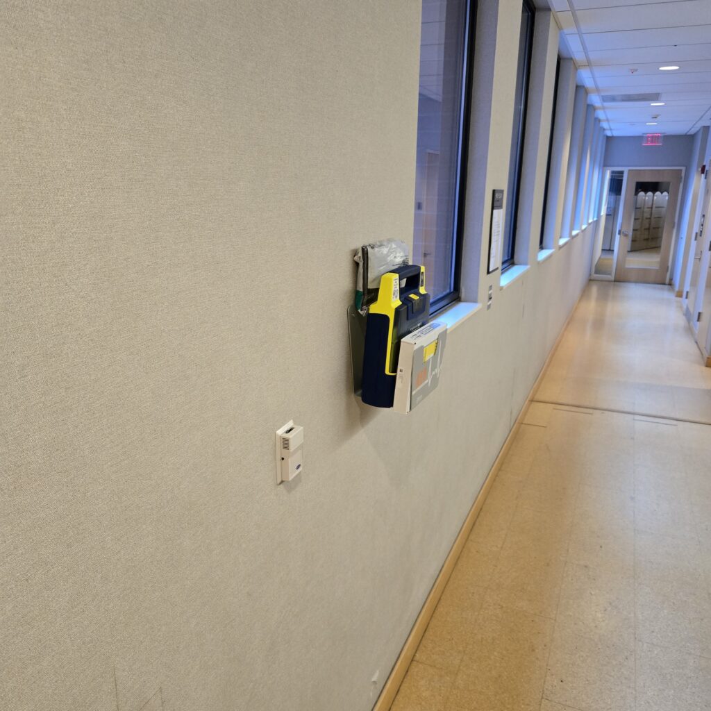 An AED mounted on the wall of a hallway with windows further along.