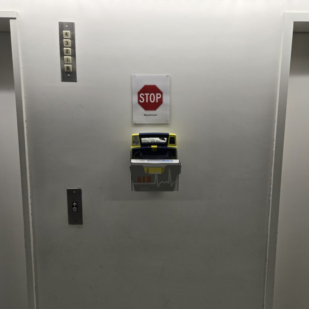 An AED mounted on a wall between elevators below a sign saying STOP.