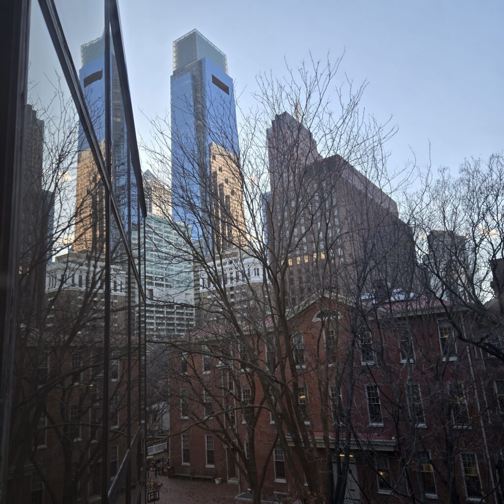 Skyscrapers and a blue sky are in the background, with a lower red brick building in the midground. A bare tree is in front of them. On the left, a window wall reflects the scene. It is a view from the Friends Center office building.