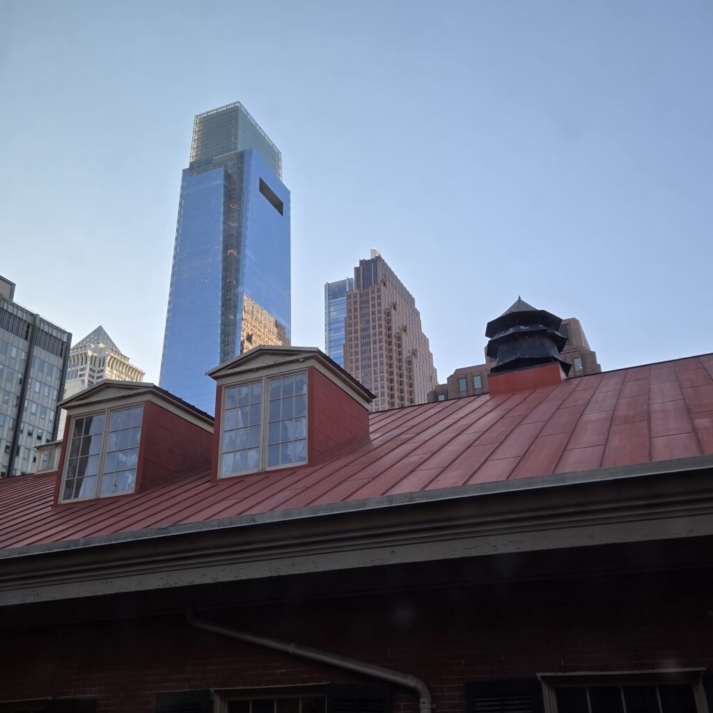 Skyscrapers and a blue sky are in the background, with a red metal roof in the foreground. It is a view from the Friends Center office building of the roof of the Race Street Quaker Meetinghouse in Center City Philly.