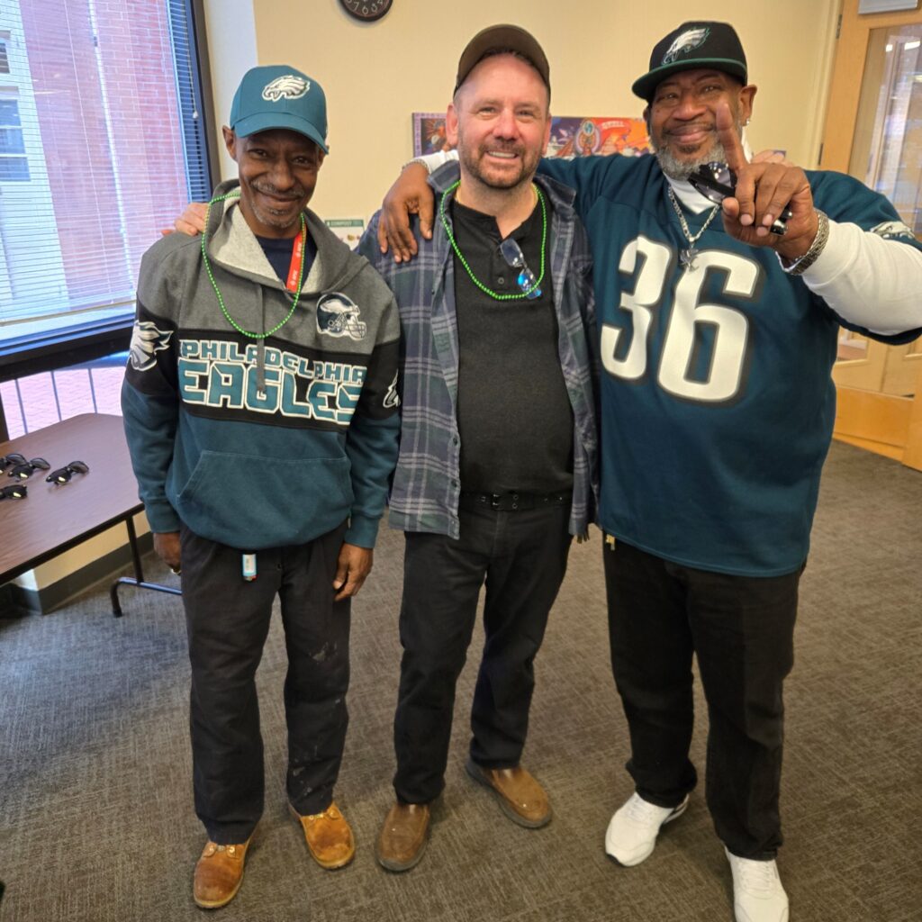 Three men smiling. The ones on left and right are wearing Eagles hats and tops.