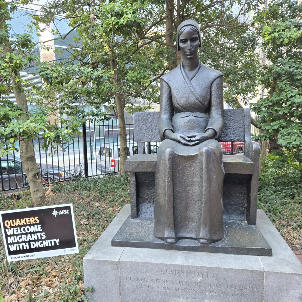 A bronze statue of a woman in plain dress with a head covering sitting on a bench. Her hands are in her lap. Her face is solemn and her gaze is directed somewhat downward. The statue is on an engraved stone plinth that says, "Mary Dyer. Quaker witness for religious freedom. Holly trees are behind her. On the left is a yard sign that says, "AFSC. Quakers welcome migrants with dignity."