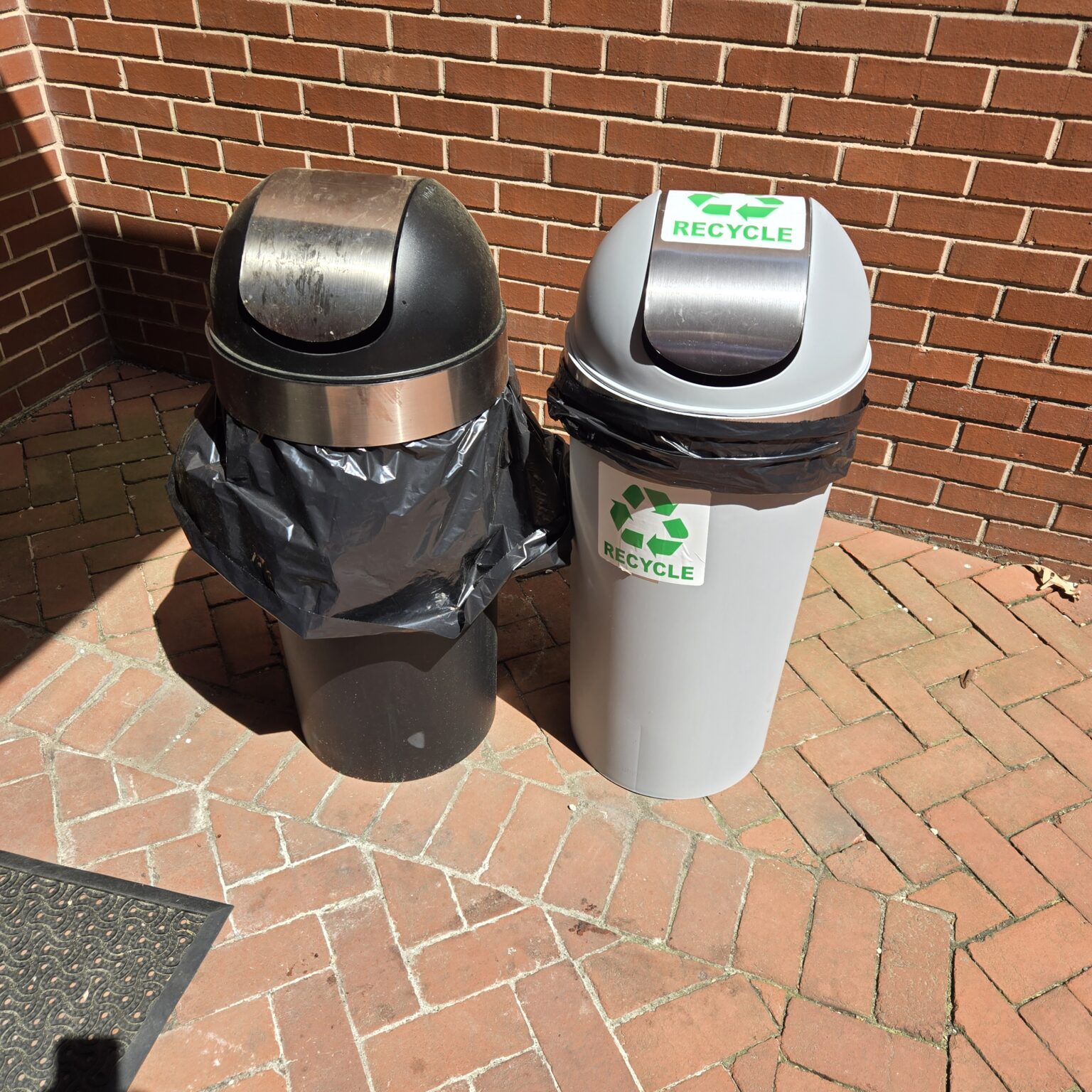 Two trash cans sit on brick pavers next to a brick wall. One is black. The other is gray and has two stickers that say Recycle and have the recycling logo consisting of three arrows twisted into a triangle shape.