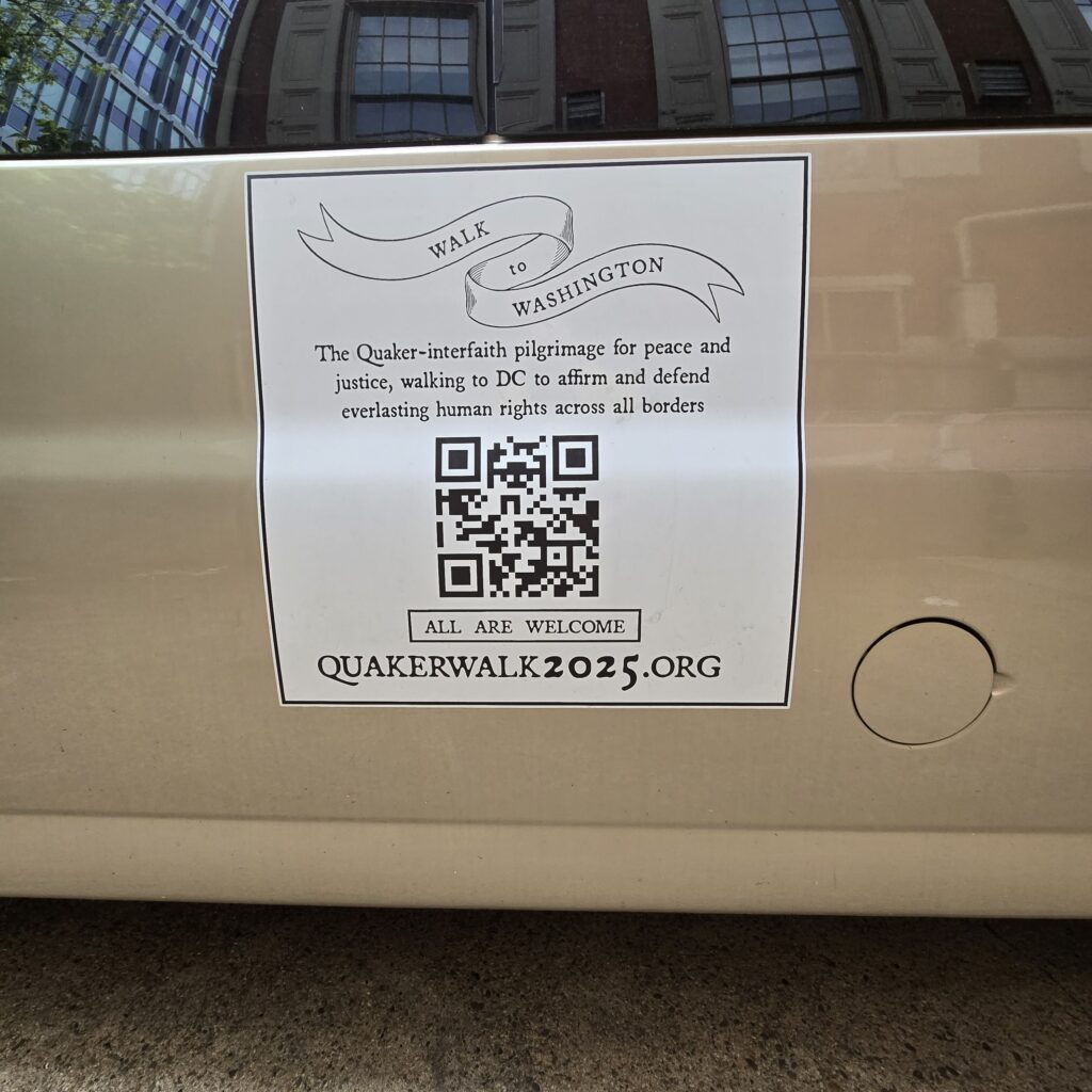The side of a van with a decal that says: "Walk to Washington. Walk to Washington. The Quaker-interfaith pilgrimage for peace & justice, walking to DC to affirm & defend everlasting human rights across all borders. ALL ARE WELCOME. Quakerwalk2025.org." The Race St. Quaker Meetinghouse's windows are reflected in the van window!