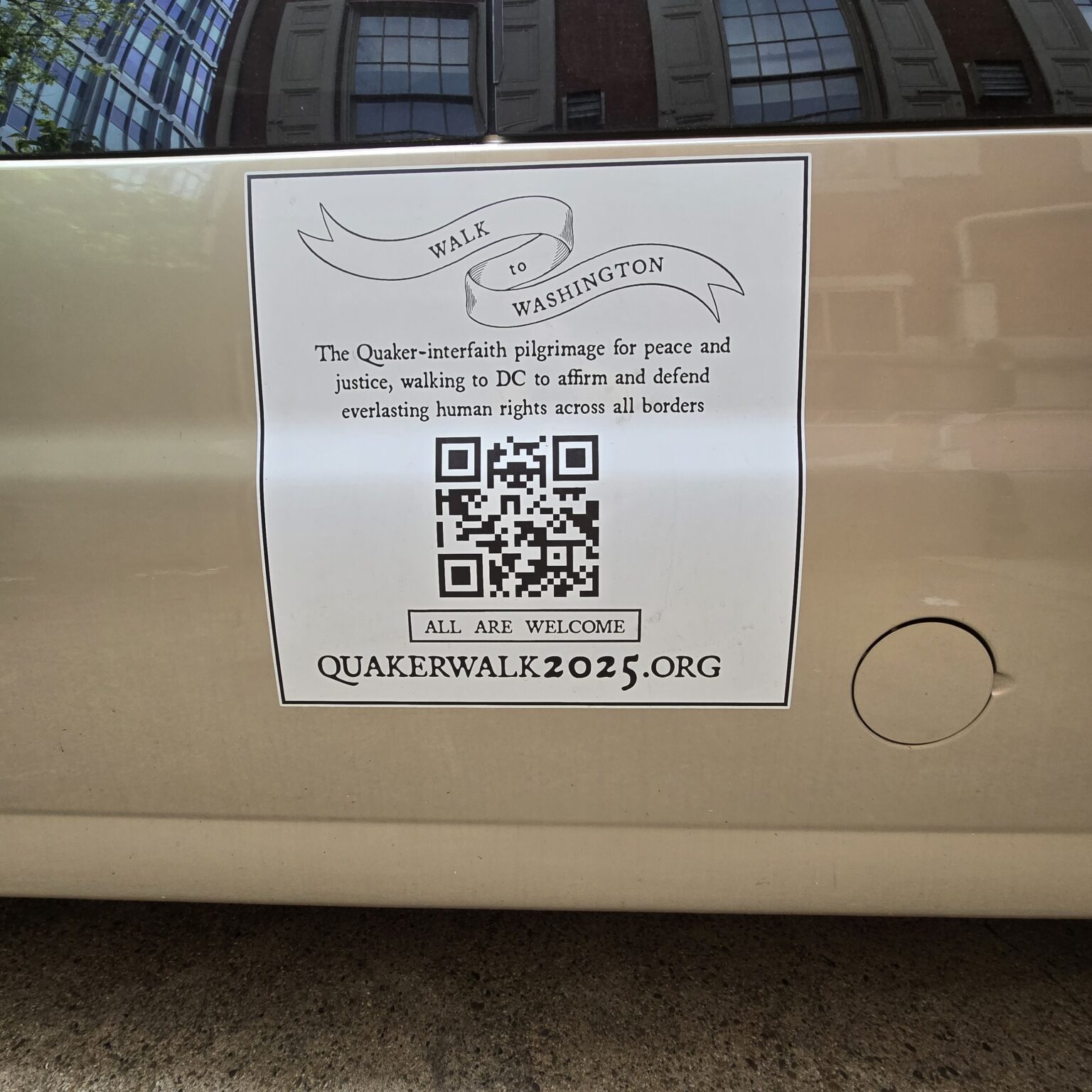 The side of a van with a decal that says: "Walk to Washington. Walk to Washington. The Quaker-interfaith pilgrimage for peace & justice, walking to DC to affirm & defend everlasting human rights across all borders. ALL ARE WELCOME. Quakerwalk2025.org." The Race St. Quaker Meetinghouse's windows are reflected in the van window!