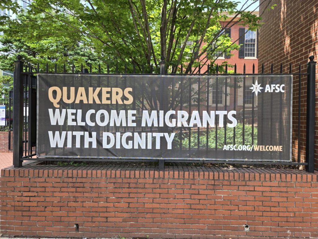Banner says,"Quakers welcome migrants with dignity. afsc.org/welcome." It is hung on a black fence above a low brick wall. In the background are green trees and the Race Street Quaker Meetinghouse.