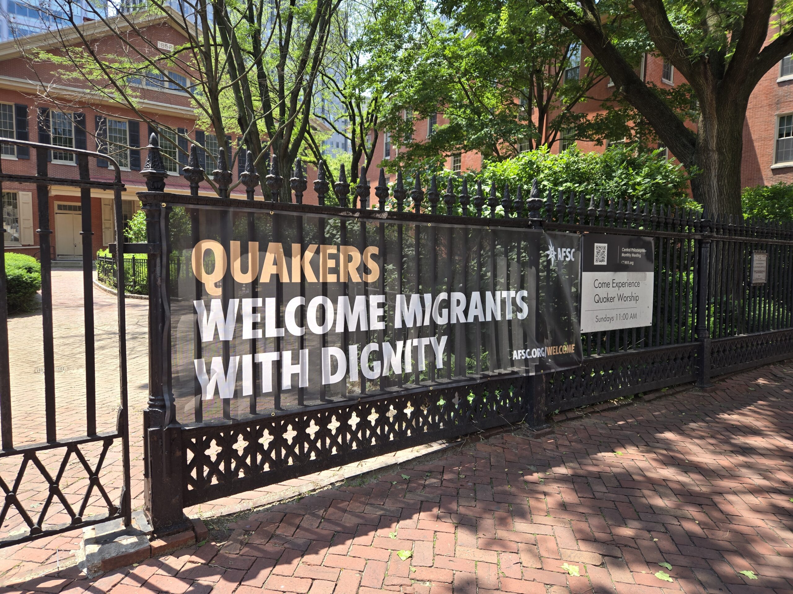 Banner says,"Quakers welcome migrants with dignity. afsc.org/welcome." It is hung on a cast iron black fence on a brick sidewalk. In the background are green trees, the Race Street Quaker Meetinghouse, and Friends Select School's STEAM Building. Another sign says, "Come experience Quaker worship, Sundays, 11 am."