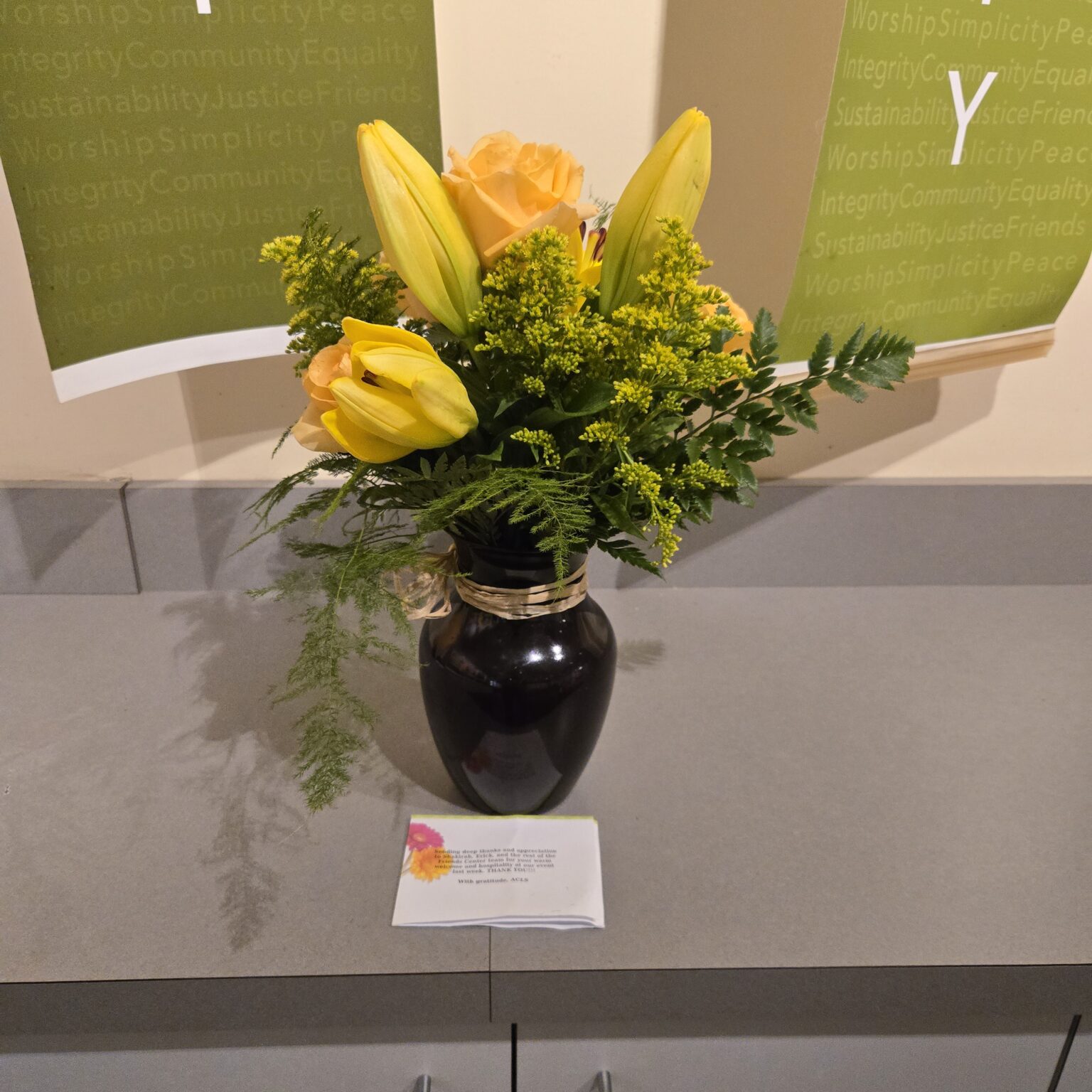 A small vase of flowers sits on a gray countertop. It has green ferns and filling. Yellow roses and lilies spring up above the greens. In the background are the bottom ends of two green banners against a yellow wall. A note is in front of the vase.
