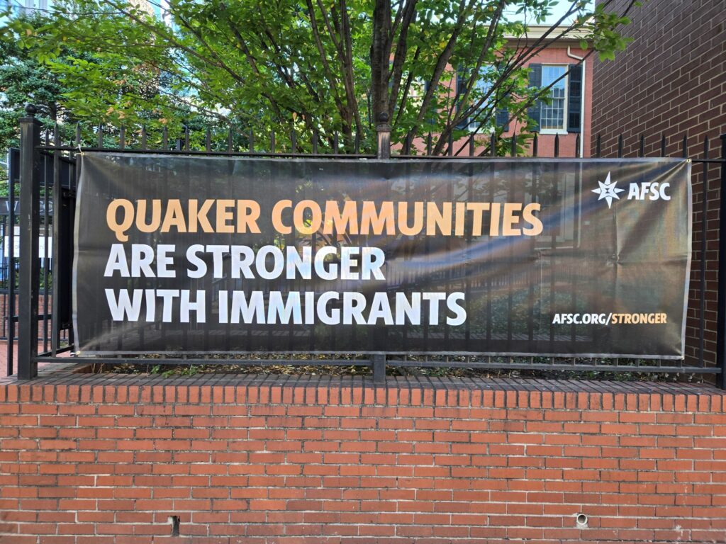 Banner on a black metal fence says, "Quaker communities are stronger with immigrants." The AFSC logo is in the upper right corner. The lower left has the url afsc.org/stronger. The fence is installed on a brick wall. In the background is a tree and the Race Street Quaker Meetinghouse. This is on 15th Street.
