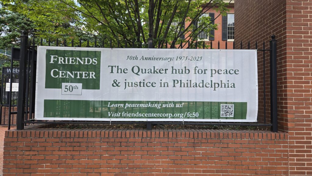 A white and green banner hangs on a black fence above a red brick wall. In the background are trees and the Race Street Quaker Meetinghouse. The banner says, Friends Center 50th. 50th anniversary 1975-2025. The Quaker hub for peace and justice in Philadelphia. Learn peacemaking with us." There is a web address and a QR code that go to the web page linked in the post.